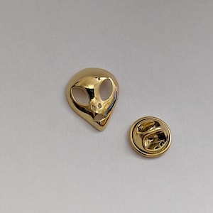 Peut inclure: Une épingle de revers en forme de tête d'extraterrestre dorée et un fermoir assorti. La tête d'extraterrestre a de grands yeux ovales et une petite bouche. Le fermoir est rond avec un fermoir.