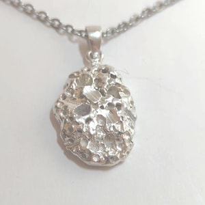 May include: Silver necklace with a textured, irregular pendant. The pendant has a rough, organic appearance, like a small rock or nugget. The delicate chain complements the pendant's design. The necklace is shown against a white background.