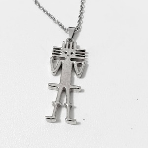 Puede incluir: Un collar de cadena de plata con un colgante que presenta una figura humana estilizada. La figura está hecha de plata y tiene un diseño simple y geométrico.