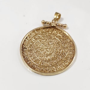 Puede incluir: Un colgante dorado con un diseño circular con detalles intrincados y una anilla en forma de anillo. El colgante recuerda a un artefacto antiguo o una moneda histórica.