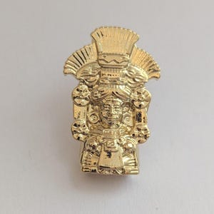 Pode incluir: Estatueta dourada de uma figura com um elaborado cocar e traços faciais detalhados. A estatueta tem uma base retangular e está sobre um fundo branco liso. O design é intrincado.