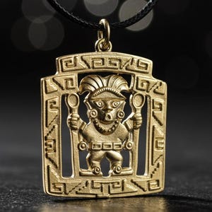 Puede incluir: Collar con colgante dorado que presenta una figura estilizada sosteniendo objetos, enmarcada en un patrón geométrico rectangular. El colgante cuelga de un cordón negro, sobre un fondo oscuro.