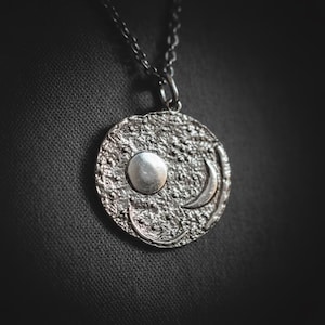 以下が含まれることがあります： テクスチャ加工された円形ペンダントが付いた、シルバーカラーのペンダントネックレス。ペンダントには、太陽と三日月のデザインが施されています。背景は暗いです。