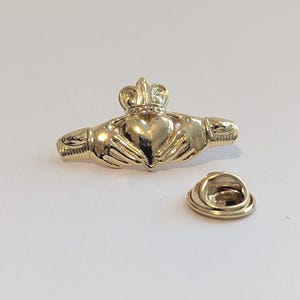 Könnte beinhalten: Goldfarbene Claddagh-Anstecknadel mit einem Herzen, das von zwei Händen gehalten und von einer Krone gekrönt wird. Das traditionelle irische Design symbolisiert Liebe, Loyalität und Freundschaft. Die Nadel hat einen separaten Verschluss.