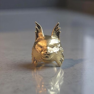 May include: A gold-toned metal ring featuring a sculpted lynx head with pointed, tufted ears and detailed facial fur textures. The ring has a polished, reflective surface that catches the light, showcasing the intricate feline features.