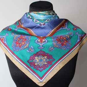 May include: Square silk scarf with a vibrant design. The scarf features a teal green center with red, yellow, and blue floral patterns. A light blue and purple border frames the design, with a tan border. The scarf is displayed on a black mannequin head.