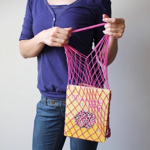 May include: A bright pink, hand-knitted net bag holding a yellow book with text and a floral design. The bag is held by a person wearing a blue shirt and jeans. The bag's open weave design is visible.