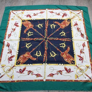 May include: A square silk scarf featuring a deep green border and a central navy blue panel. The design includes illustrations of brown saddles, gold horseshoes, golden chains, hunting horns, and red bird motifs arranged in a symmetrical pattern.