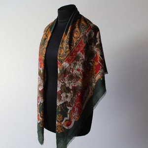 Può includere: Un foulard quadrato con bordo verde scuro e un motivo centrale di fiori e volute ornamentali nei toni del rosso, marrone e oro. Il tessuto ha una consistenza morbida con bordi sfrangiati visibili, drappeggiato su un manichino nero.