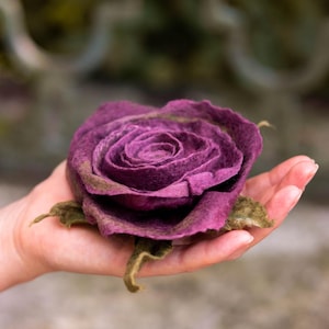 Può includere: Una rosa in feltro fatta a mano, nei toni del viola e del verde. Il fiore è tenuto in una mano, mostrando il suo design dettagliato dei petali e la forma realistica. La rosa in feltro è una creazione artistica unica.