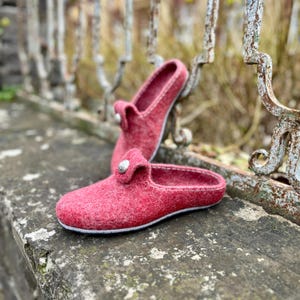 May include: A pair of red felt slippers with a button detail. The slippers are a muted red color with a white stitched sole. One slipper is in the foreground, and the other is in the background, resting on a stone surface.