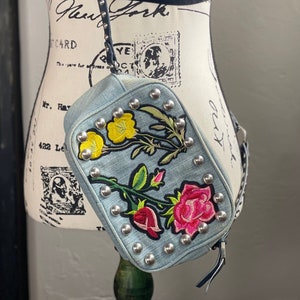 May include: A light blue denim crossbody bag with silver studs and floral embroidery. The bag has a black strap and a rectangular shape.