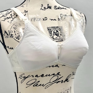 May include: A white bra with adjustable straps and a delicate lace trim. The bra is displayed on a mannequin. The bra has a classic design with a small bow detail at the center.