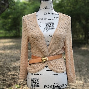 May include: A beige crocheted cardigan with a V-neck and long sleeves. The cardigan is belted with a brown leather belt with a gold buckle. The cardigan is on a white mannequin.