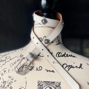 May include: White leather belt with silver-tone buckle and decorative studs. The belt is wrapped around a mannequin. The buckle and studs have a floral design. The belt is approximately 2.5 cm wide.