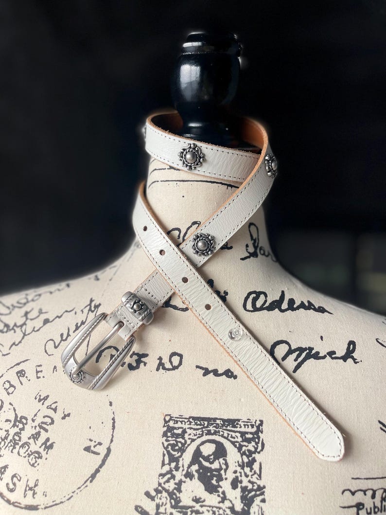 May include: White leather belt with silver-tone buckle and decorative studs. The belt is wrapped around a mannequin. The buckle and studs have a floral design. The belt is approximately 2.5 cm wide.