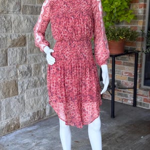 Pode incluir: Vestido coral com padr&atilde;o floral, exibido em um manequim. O vestido apresenta mangas compridas e transparentes, um corpete franzido e uma saia plissada. Possui decote redondo e cintura ajustada. A bainha &eacute; assim&eacute;trica. O vestido tem cerca de 100 cm de comprimento.