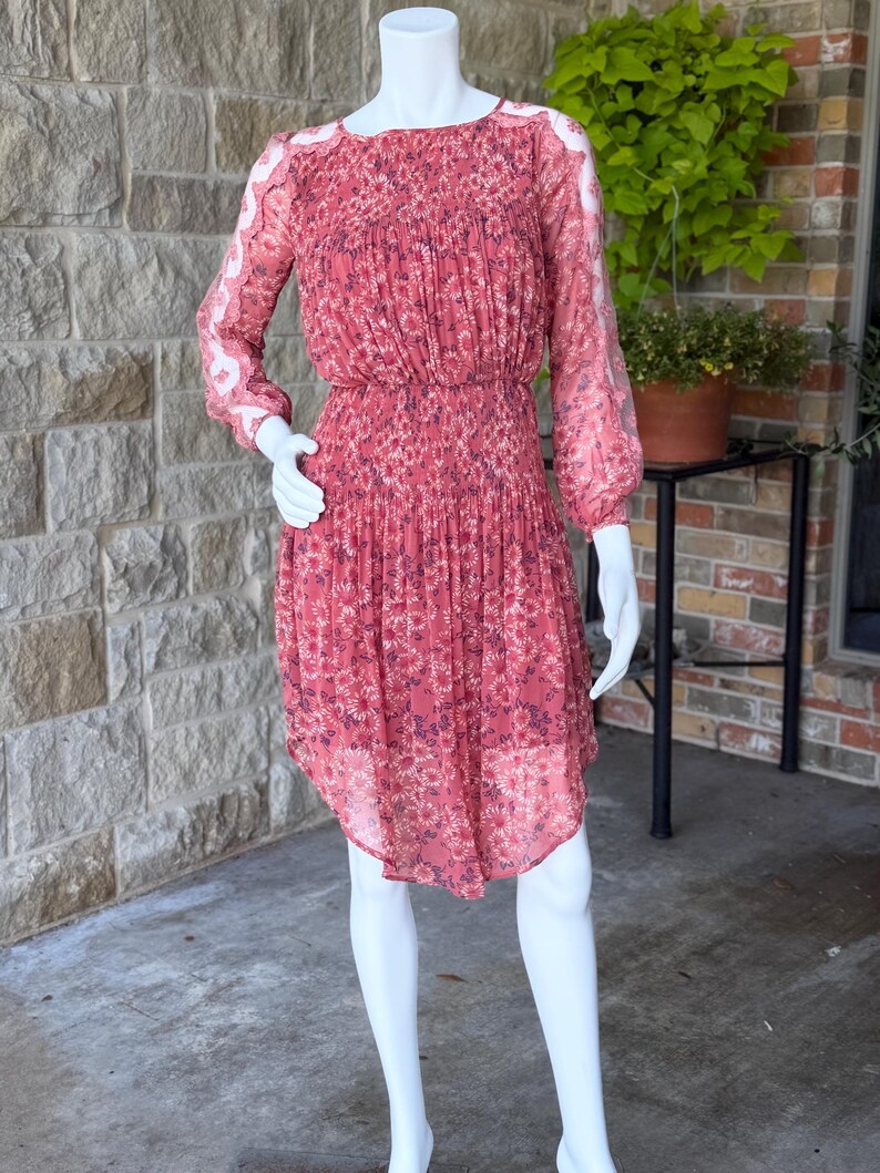 Pode incluir: Vestido coral com padr&atilde;o floral, exibido em um manequim. O vestido apresenta mangas compridas e transparentes, um corpete franzido e uma saia plissada. Possui decote redondo e cintura ajustada. A bainha &eacute; assim&eacute;trica. O vestido tem cerca de 100 cm de comprimento.