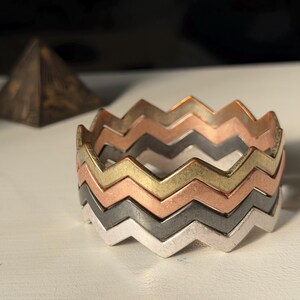 May include: A stack of four interlocking metal bracelets featuring a repeating zigzag or chevron pattern. The bangles are finished in distinct metallic tones, including silver, dark gray, copper, and gold, creating a layered, multi-colored effect.