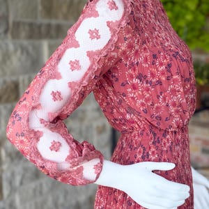Pode incluir: Vestido de manga comprida com estampa floral em tons de vermelho e rosa. As mangas apresentam um design de renda exclusivo com uma borda ondulada. O vestido &eacute; exibido em um manequim.