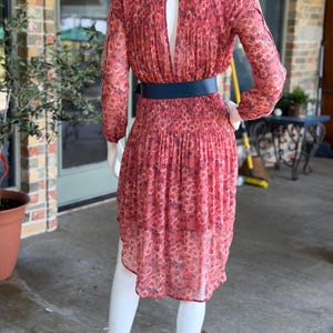 Pode incluir: Vestido coral com padr&atilde;o floral e mangas compridas. O vestido apresenta um decote nas costas e &eacute; cintado na cintura com um cinto azul marinho. O vestido &eacute; exibido num manequim.