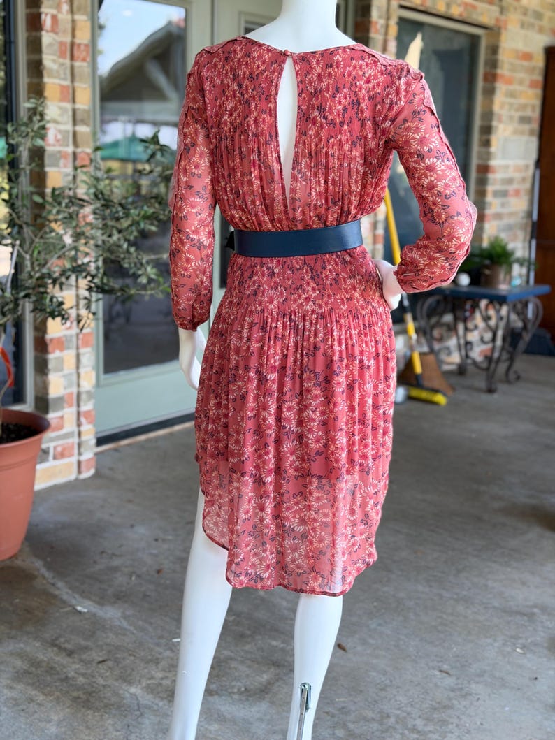 Pode incluir: Vestido coral com padr&atilde;o floral e mangas compridas. O vestido apresenta um decote nas costas e &eacute; cintado na cintura com um cinto azul marinho. O vestido &eacute; exibido num manequim.