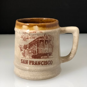 May include: A beige ceramic mug with a brown rim and handle. The mug features a brown illustration of a San Francisco cable car and the words "SAN FRANCISCO" below the image.