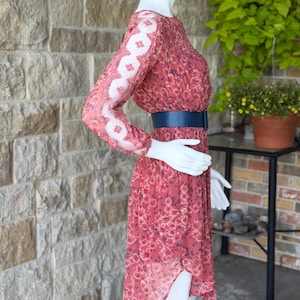 Pode incluir: Vestido coral com estampa floral e mangas compridas. O vestido apresenta um cinto azul e uma saia fluida. As mangas t&ecirc;m um design de renda floral branca. O vestido &eacute; exibido em um manequim.