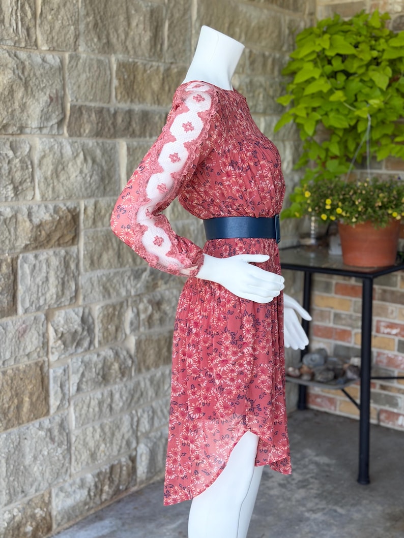 Pode incluir: Vestido coral com estampa floral e mangas compridas. O vestido apresenta um cinto azul e uma saia fluida. As mangas t&ecirc;m um design de renda floral branca. O vestido &eacute; exibido em um manequim.