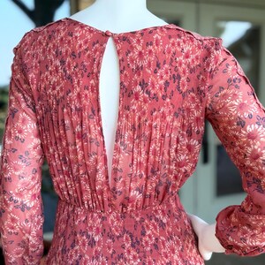 Pode incluir: Vestido coral de manga comprida com padr&atilde;o floral. O vestido apresenta um detalhe nas costas em forma de fechadura e detalhes plissados. O tecido parece leve e fluido, com cintura franzida. O vestido &eacute; exibido num manequim.