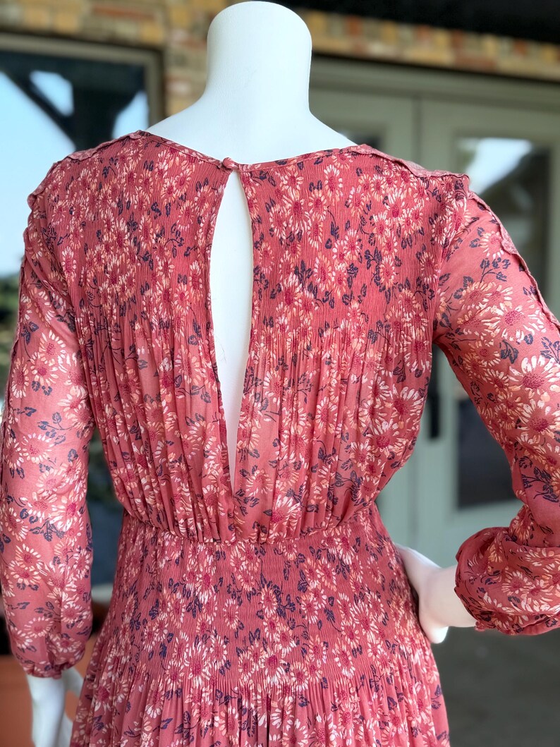 Pode incluir: Vestido coral de manga comprida com padr&atilde;o floral. O vestido apresenta um detalhe nas costas em forma de fechadura e detalhes plissados. O tecido parece leve e fluido, com cintura franzida. O vestido &eacute; exibido num manequim.
