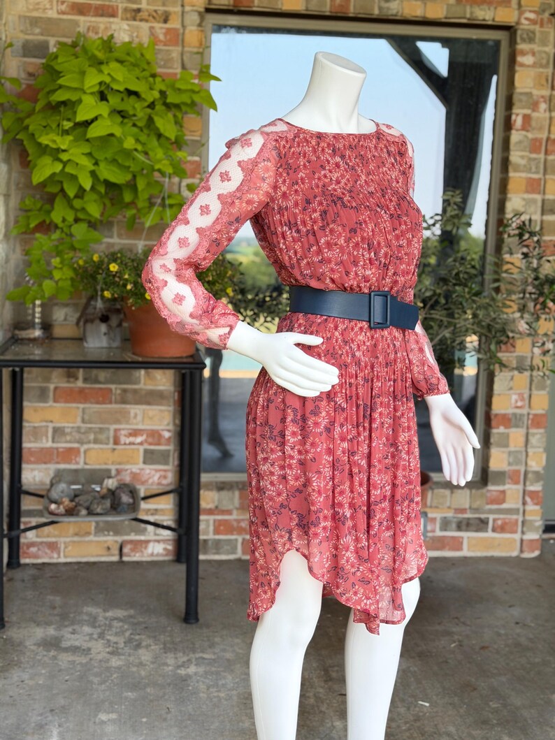 Pode incluir: Um vestido vermelho com estampa floral, mangas compridas e cinto azul. O vestido tem um corpete plissado e uma saia fluida. As mangas t&ecirc;m um detalhe transparente semelhante a renda. O vestido &eacute; exibido em um manequim.