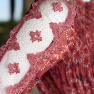Pode incluir: Close-up de uma manga de vestido cor de ferrugem com estampa floral. A manga apresenta pain&eacute;is de renda branca com detalhes florais bordados e bordas onduladas em uma cor de ferrugem combinando. O vestido parece ser feito de um tecido leve e fluido.