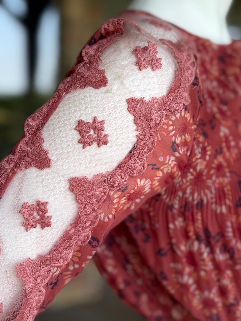Pode incluir: Close-up de uma manga de vestido cor de ferrugem com estampa floral. A manga apresenta pain&eacute;is de renda branca com detalhes florais bordados e bordas onduladas em uma cor de ferrugem combinando. O vestido parece ser feito de um tecido leve e fluido.
