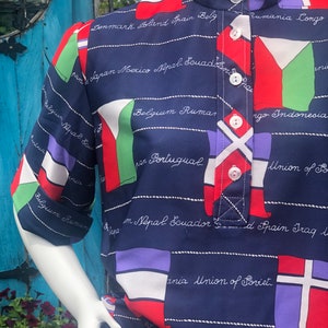May include: A navy blue button-down shirt with a pattern of flags from different countries. The shirt has a white collar and a button-up closure. The flags are in various colors, including red, white, green, blue, and yellow. The shirt has a text pattern that includes the names of countries, such as "Denmark", "Spain", "Belgium", "Rumania", "Congo", "Japan", "Mexico", "Nepal", "Ecuador", "Portugal", "Indonesia", and "Union of Soviet".