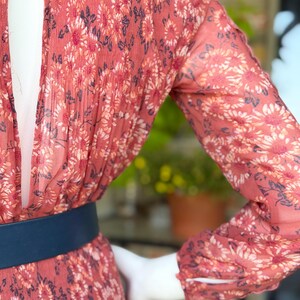 Pode incluir: Vestido com estampa floral em tons de coral, branco e azul marinho. O vestido apresenta mangas compridas, cintura marcada com cinto azul marinho e decote em forma de fechadura. O tecido parece leve e fluido.