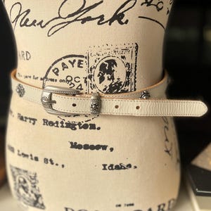 May include: Cream leather belt with a silver buckle and decorative studs. The belt is displayed on a mannequin. The buckle is rectangular with a floral design. The belt has a classic, Western-inspired style.