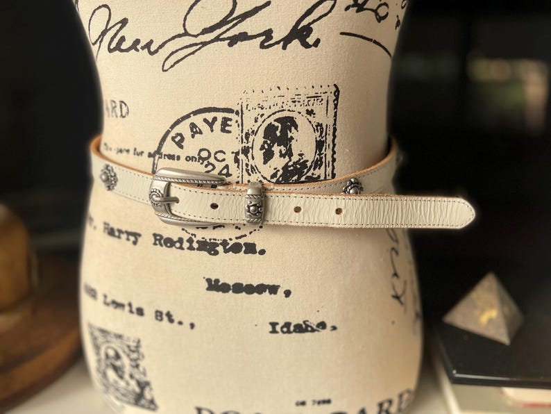 May include: Cream leather belt with a silver buckle and decorative studs. The belt is displayed on a mannequin. The buckle is rectangular with a floral design. The belt has a classic, Western-inspired style.