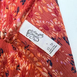 Pode incluir: Close-up de uma pe&ccedil;a de roupa cor de ferrugem com um padr&atilde;o floral em tons de laranja, rosa e azul marinho. Uma etiqueta branca com o log&oacute;tipo "Free People" e o tamanho "XS TP" &eacute; vis&iacute;vel. O tecido parece leve e fluido.