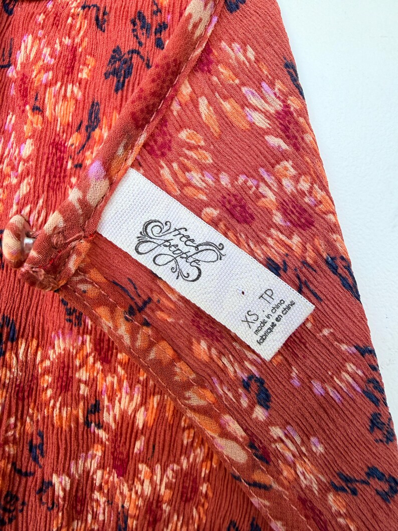 Pode incluir: Close-up de uma pe&ccedil;a de roupa cor de ferrugem com um padr&atilde;o floral em tons de laranja, rosa e azul marinho. Uma etiqueta branca com o log&oacute;tipo "Free People" e o tamanho "XS TP" &eacute; vis&iacute;vel. O tecido parece leve e fluido.