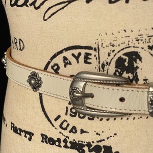 May include: Cream leather belt with a silver-tone buckle and decorative metal studs. The belt has a classic design with detailed embellishments, suitable for adding a touch of style to any outfit.