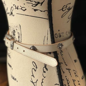 May include: Cream leather belt with silver-tone metal buckle and decorative studs. The belt is wrapped around a mannequin. The mannequin is covered in script and stamps.