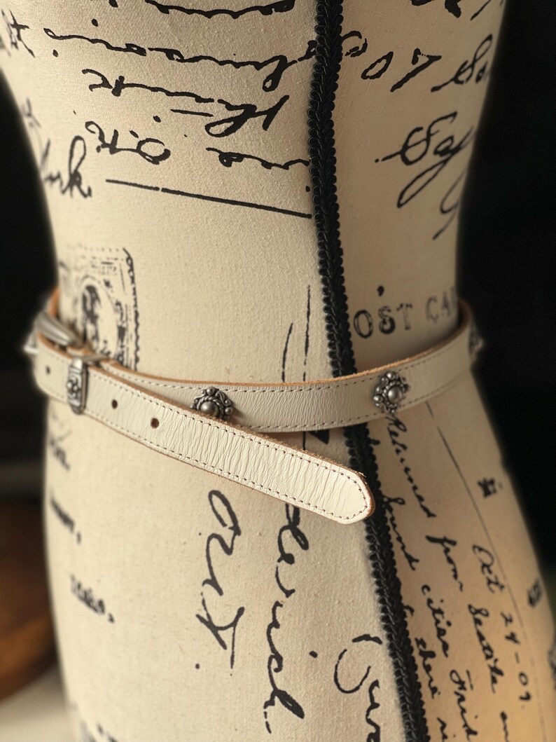 May include: Cream leather belt with silver-tone metal buckle and decorative studs. The belt is wrapped around a mannequin. The mannequin is covered in script and stamps.