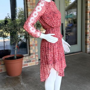 Pode incluir: Vestido vermelho com estampa floral em manequim. O vestido tem mangas compridas com um padr&atilde;o de renda floral branca transparente na parte superior dos bra&ccedil;os. A saia &eacute; plissada e assim&eacute;trica, com bainha frontal mais curta.