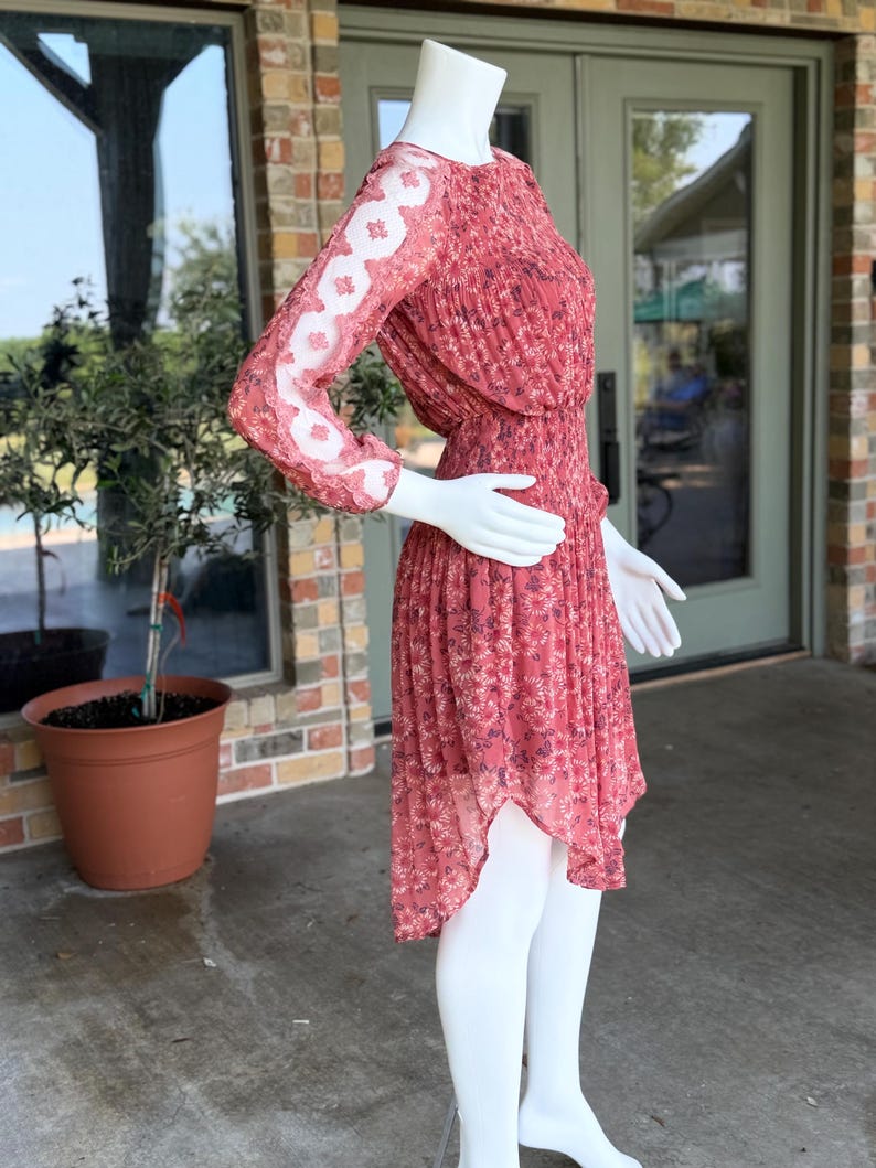 Pode incluir: Vestido vermelho com estampa floral em manequim. O vestido tem mangas compridas com um padr&atilde;o de renda floral branca transparente na parte superior dos bra&ccedil;os. A saia &eacute; plissada e assim&eacute;trica, com bainha frontal mais curta.