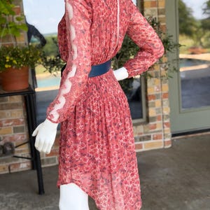 Pode incluir: Um vestido vermelho com estampa floral, mangas compridas e cinto azul. O vestido tem uma saia plissada e mangas transparentes com detalhes em renda. O vestido &eacute; exibido em um manequim.