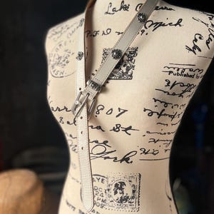 May include: A pale grey leather belt with silver metal buckles and floral embellishments. The belt is displayed on a mannequin with a vintage design, highlighting its details and craftsmanship.