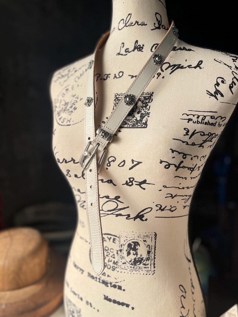 May include: A pale grey leather belt with silver metal buckles and floral embellishments. The belt is displayed on a mannequin with a vintage design, highlighting its details and craftsmanship.