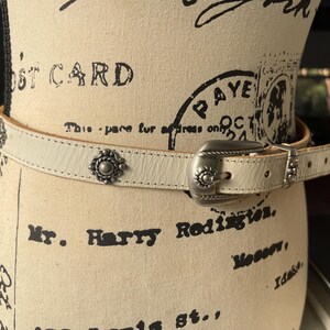 May include: Cream leather belt with a silver-tone buckle and decorative studs. The belt is displayed on a mannequin with vintage text and stamps. A fashionable accessory.