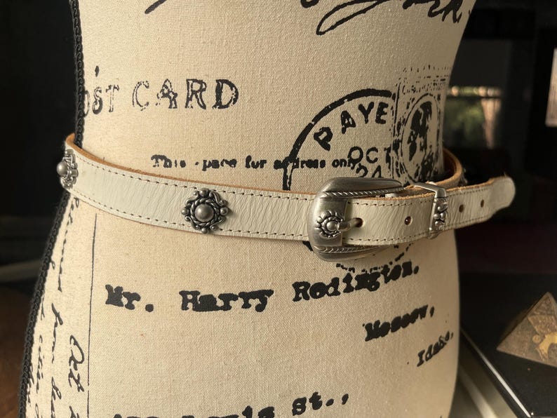 May include: Cream leather belt with a silver-tone buckle and decorative studs. The belt is displayed on a mannequin with vintage text and stamps. A fashionable accessory.